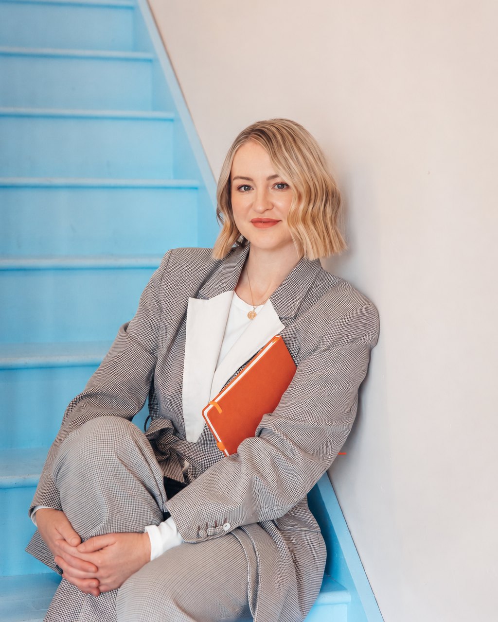 Melanie Jolly, founder of The Perinatal Workplace Consultancy, sitting on a blue staircase holding an orange notebook